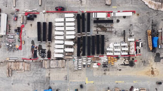 Large construction site with rows of white prefab units, black pipe segments, lumber stacks and heavy equipment spread across concrete yard in Lancaster, Pennsylvania, USA. Aerial drone top down.