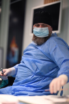 Bearded Surgeon In Surgical Gown And Mask Reaching During Procedure In Hospital Operating Room