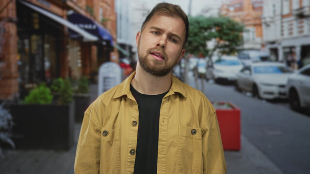Man tilts head, beard visible on a city street with parked cars and planters; pensive quiet thought.