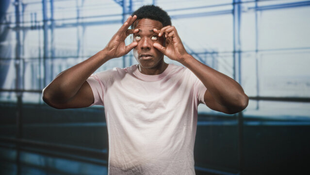 Black man pulls down lower eyelids with fingers and rolls eyes in airport terminal wearing pink tshirt and looking upward; tired travel delay.
