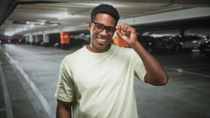 Fototapeta premium Man adjusting glasses with hand in a parking garage building, smiling and looking at camera, wearing light t shirt and visible forearm; confidence.