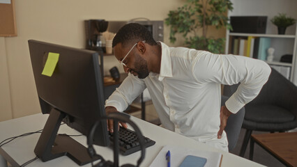 Man hold lower back for backache at office while typing at desktop keyboard and looking at monitor,...