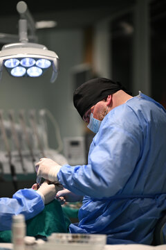 Male Dentist Performing Dental Surgery In Clinic Wearing Surgical Gown Mask And Head Cover