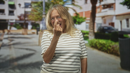 Fototapeta premium Senior blonde woman winks and beckons with fingers, bare hand forward, wearing striped shirt in a sunlit pedestrian street with palm trees; playful teasing.