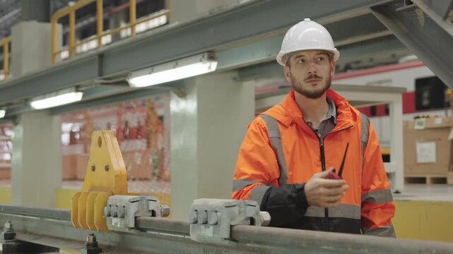 Focused engineer inspects metal beam within dimly lit warehouse using orange safety jacket and white hard hat during structural assessment work for electrical train maintenance.