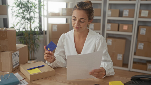Woman holds creditcard and reads a shipping invoice at a desk surrounded by parcels in a building; focused small business.