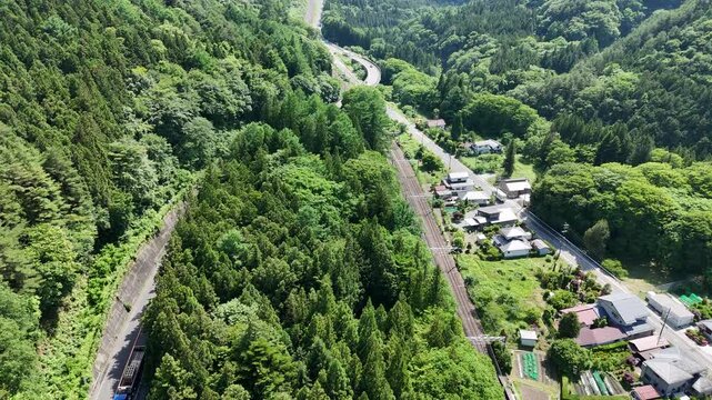 Aerial view shows winding roads through lush green forests. Small village nestled among towering trees appears serene. Village homes, roads, and railway lines follow natural contours