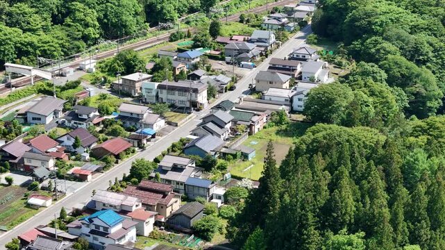 Aerial view of quiet village nestled among green hills. Road runs through homes with varied roof colors. Rail track runs parallel to the village street. Trees surround the settlement