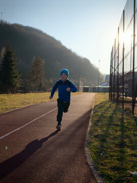 Young Boy Running on Outdoor Track at Sunset in Blue Jacket and Beanie