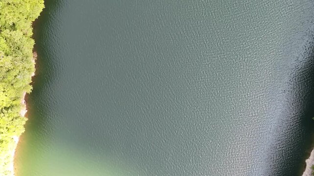 Aerial view captures calm lake surface. Green forest edge frames the water. Sunlight glints gently on the ripples. Serene landscape offers natural beauty. Ideal for tranquil nature or travel visuals