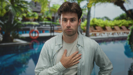 Fototapeta premium Young hispanic man with hand on chest at resort poolside, casual shirt and earnest expression; concern sincerity.