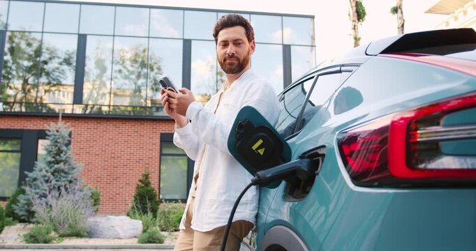 Adult man using mobile app to charge an electric car at a parking lot outdoors.