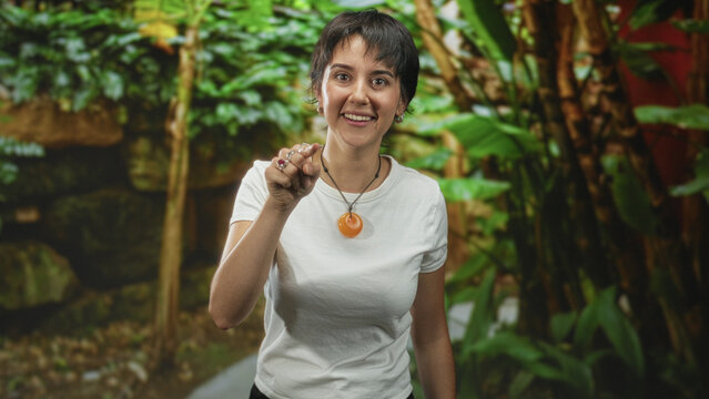 Woman points finger while wearing orange pendant necklace and white tee on a botanical garden path; assertive instruction.