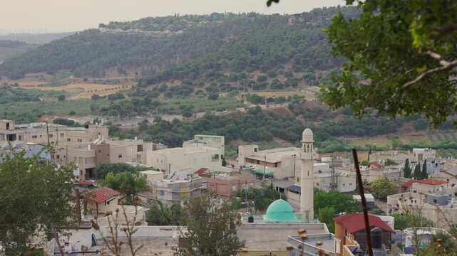Soft light bathes quiet Arab village nestled in green hills. White buildings cluster close, red roofs peek above. Minarets rise tall, domes gleam softly under sky in Israel northern region