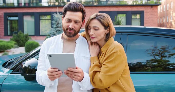 Portrait of young couple planning travel route on tablet near charging electric car in parking lot, outdoor