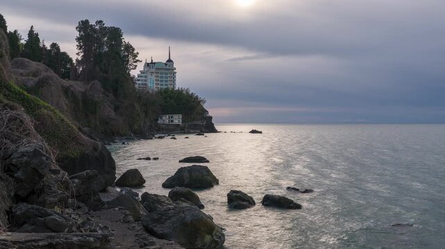 Time-lapse video of the Castello Mare resort hotel situated on a dramatic cliffside overlooking the Black Sea in Tsikhisdziri, Georgia, with clouds moving and sunlight reflecting on the water surface.