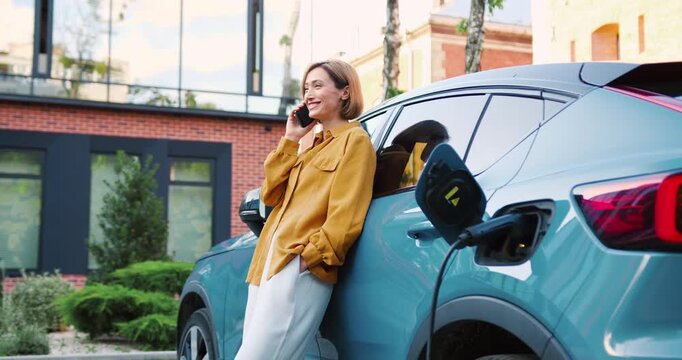 Smiling woman talking on phone near electric car connected to charging station in city parking lot, outdoor