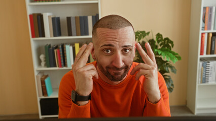Young man in orange sweater touching temples with fingers while seated at computer amid bookshelves in building study  focused concentration. © Krakenimages.com