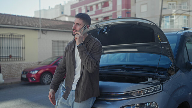 Man holding phone to ear beside car with open hood on street, leaning on bumper and looking up; roadside vehicle trouble concern.