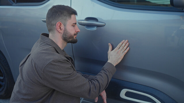 Young hispanic man kneels and touches a dent on a gray car door with bare hand while inspecting panel on street; concern.