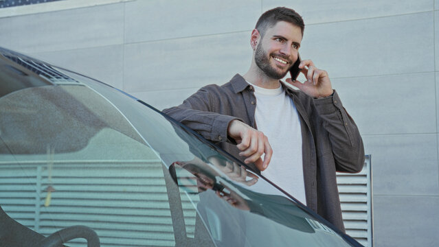 Man leaning on car holding smartphone to ear in front of building, casually resting arm on roof and smiling while talking on phone; confidence.