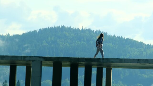 Young woman with smartphone walks away from friend along abandoned building top. Female tourists observe distant mountains from uncompleted hotel