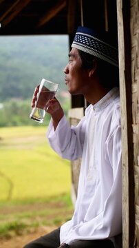 An Indonesian Muslim man drinking a glass of water on his wooden porch with scenic yellow rice field background in a rural Asian village.