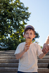 Fototapeta premium A young boy in a gray cap bites into a nearly empty waffle cone while holding an adult's hand on sunny outdoor steps surrounded by green trees and a clear blue sky