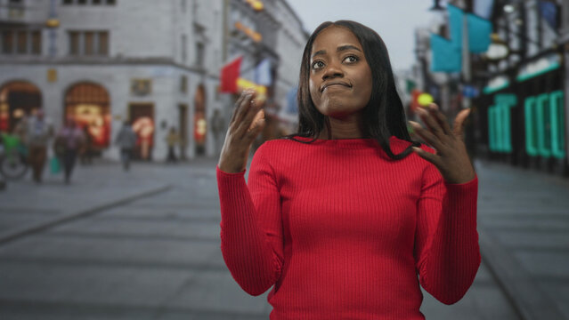 Woman in red sweater shrugs with hands raised, flipping fingers near face and pursing lips on a busy pedestrian street lined with shops and neon signs; indifference dismissal.
