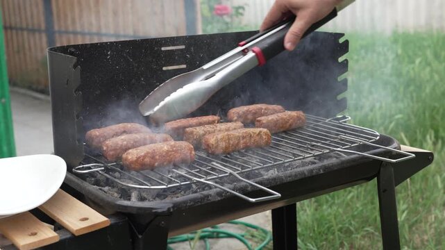 Close up of a chef using metal kitchen tongs to turn and flip traditional Romanian mici minced meat rolls cooking on a smoking charcoal barbecue grill.