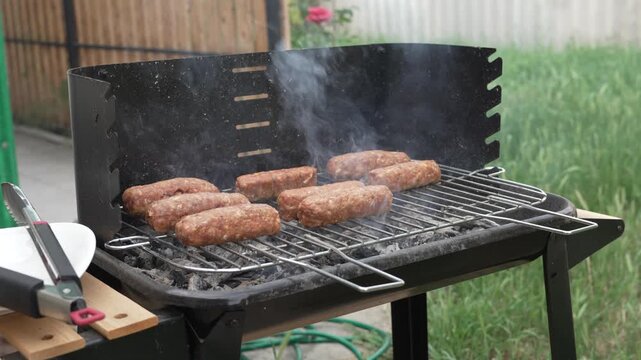 Skinless minced meat sausages, known as traditional Romanian mici or mititei, cooking on a hot wire rack over a charcoal barbecue grill with light smoke rising in a backyard.