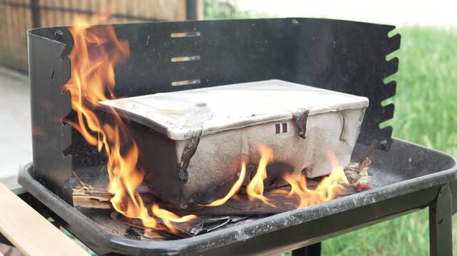 Close up of a molded paper pulp container catching fire and burning down with active orange flames and light smoke on an outdoor charcoal barbecue grill.