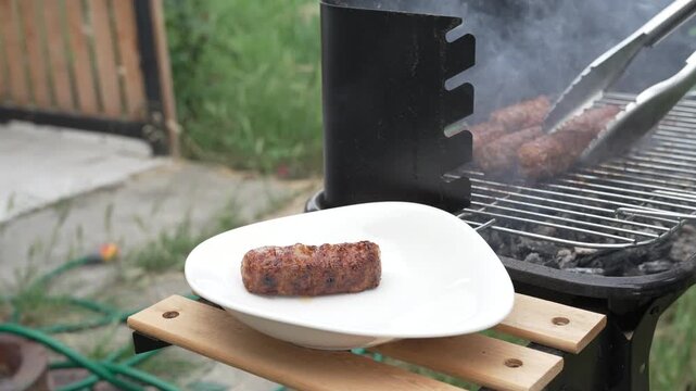 Close up of a hand using metal kitchen tongs to serve hot, steaming minced meat rolls from an outdoor barbecue grill onto a white ceramic plate in a backyard.