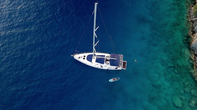 Aerial view of a white sailing yacht anchored in clear turquoise and deep blue sea, with a paddleboard nearby and visible underwater coastline, creating a peaceful Mediterranean travel stock scene.
