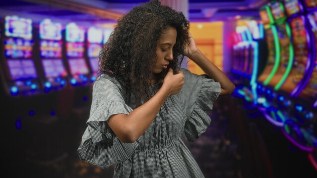 Young woman touching her neck and tucking curly hair beside glowing slot machines in a casino building; anxiety reflection.