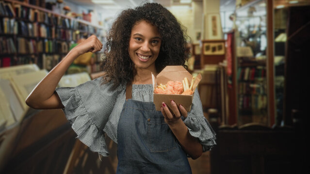 Young woman in apron holds takeaway box of fried chicken and fries, smiling and flexing arm in a bookstore building; pride.