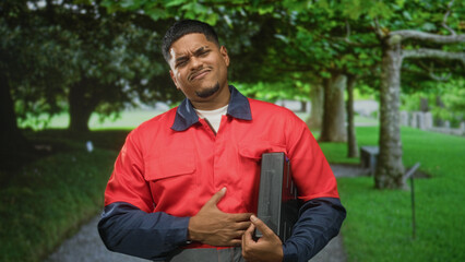 Fototapeta premium Hispanic man in red work uniform clutching chest while holding toolbox on a tree lined path in forest; pain duty resilience.