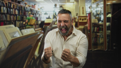 Man with clenched fists shouting in a library among bookshelves and records, grey hair and beard, fists raised in celebration  victory joy relief. © Krakenimages.com