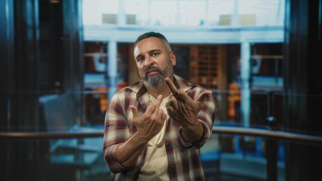 Man showing middle fingers with bare hands in building atrium near ship railing, flannel shirt and beard visible; defiance rebellion.