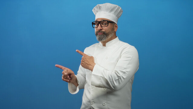 Man chef in white uniform and hat pointing with hand in studio against vivid blue backdrop; confidence skill craft.