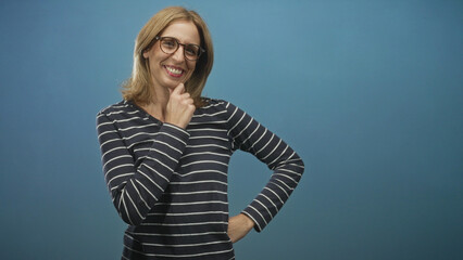 Fototapeta premium Woman wearing glasses and striped top, blonde, finger to chin and hand on hip while smiling in studio; friendly confidence.