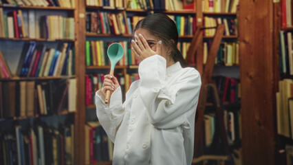 Young woman chef in white coat holds turquoise ladle and covers mouth with hand while smiling amid bookshelves in a library building  playful joy. © Krakenimages.com
