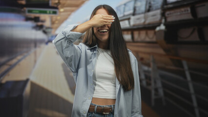 Fototapeta premium Young woman with long hair and white tee covers eyes with hand while laughing on a train platform in a building; playful joy.