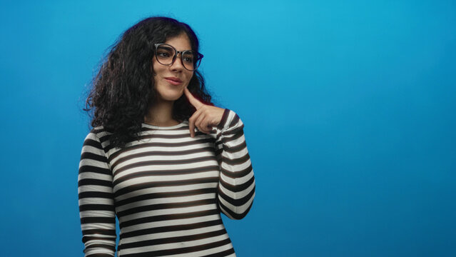 Young arab woman points index fingers to cheeks in bright blue studio wearing black and white striped shirt and oversized glasses; playful curiosity.