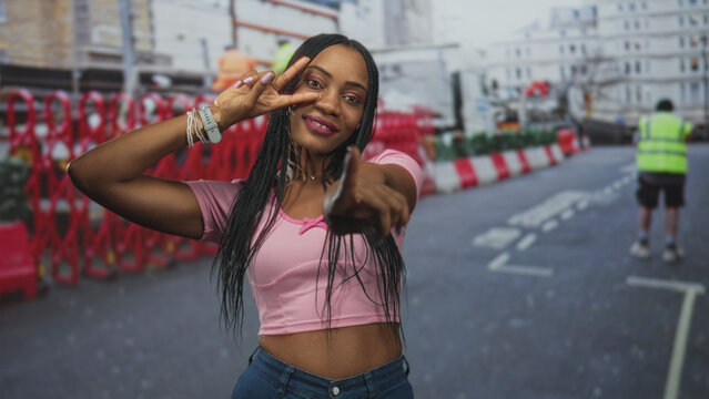 Woman pointing finger and making peace sign with bare midriff on a busy street with construction barriers and traffic cones; playful confidence.