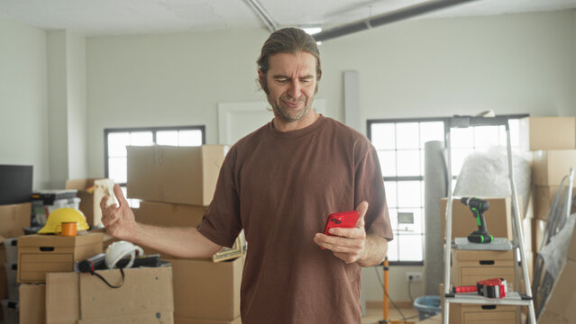 Man holding red smartphone, looking at phone and gesturing amid packed moving boxes inside a building; moving frustration.