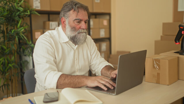 Man with beard covers face with hands at desk with laptop and parcels in a packing area in a building; small business worry.