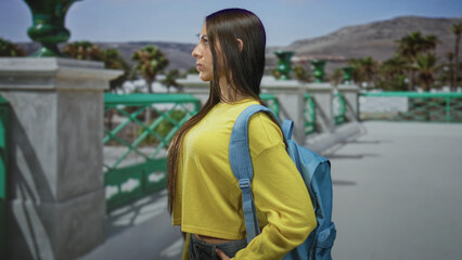 Obraz premium Young woman student in yellow sweater with blue backpack stands in profile, hand in pocket, on a street promenade by the sea; contemplation.