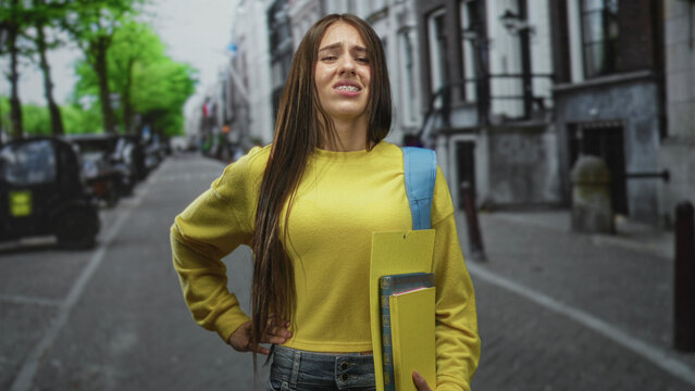 Teen student girl wearing yellow sweater and blue backpack strap, holding yellow folder and book, hand on stomach with pained wince on urban street; discomfort.