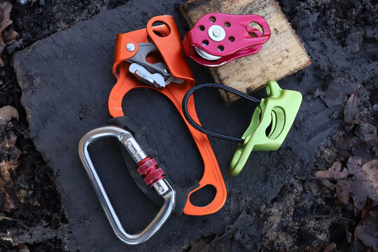 Top view flat lay of professional colorful climbing gear setup on dark textured stone background including metal carabiner, orange hand ascender rope clamp, red pulley and green belay device